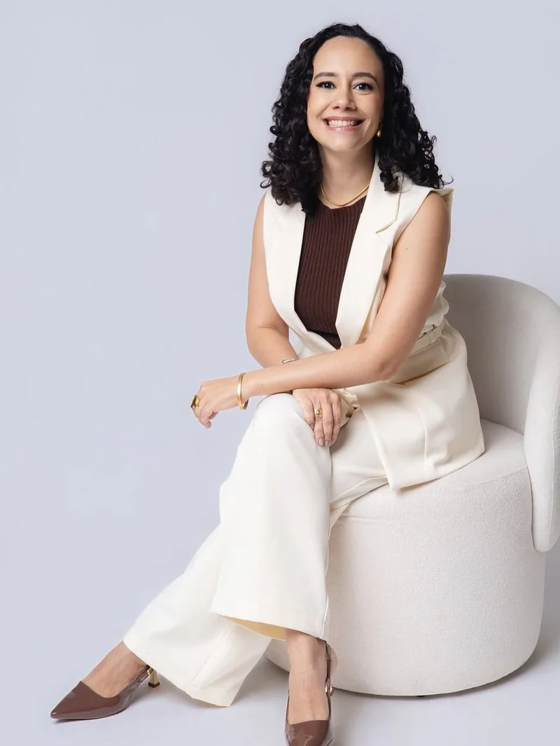 Dra. Sara Cristina sentada em poltrona, sorrindo, com blazer branco sobre blusa marrom e calça off-white
